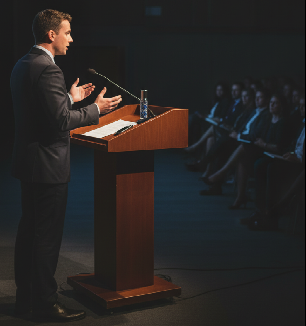 a man standing at a poteum in a suit in front of an audience giving a speech the clear glass rollerbottle blend, candorVIII sitting on poteum in front of him roller bottle infused with lapis lazuli the blend representing confidence with assertive speech, clarity in speaking your truth 