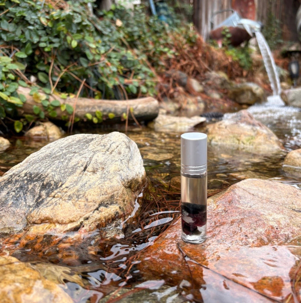 10ml clear glass bottle with silver cap the blend quell inside for used for bug bite relief infused with garnet obsidian & clear quartz sitting on rocks with a river & waterfall behind  