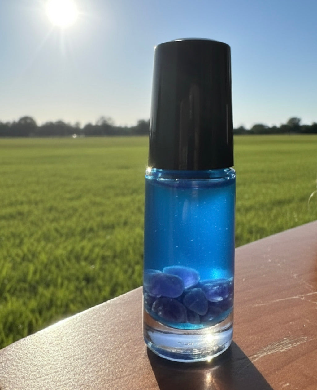 5 ml blue gradient glass bottle with black cap sitting on wood with sn shining above & a grass area behind to represent the blend solace infused with amethyst & rose quartz inside the roller bottle a blend to encourage calm & balanced emotional response instead of angry reactions 